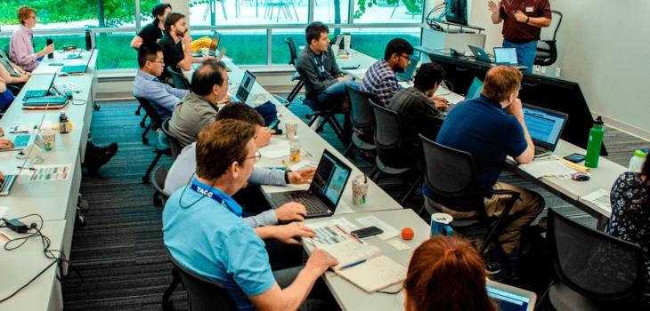 Several students sitting at long desks with laptops being instructed by a man standing at the front of the class.